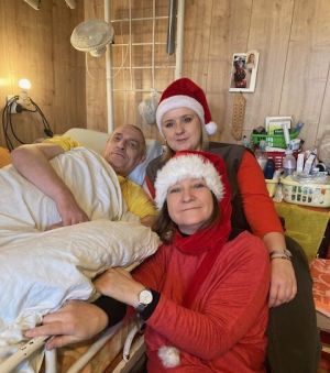 mężczyzna na łóżku rehabilitacyjnym z dwiema kobietami w czapkach świątecznych